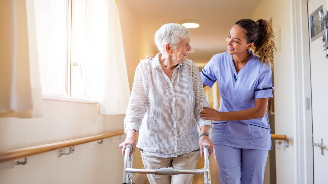 Une aide-soignante accompagne un résident âgé en déambulateur dans le couloir d'un EHPAD © SeniorActu