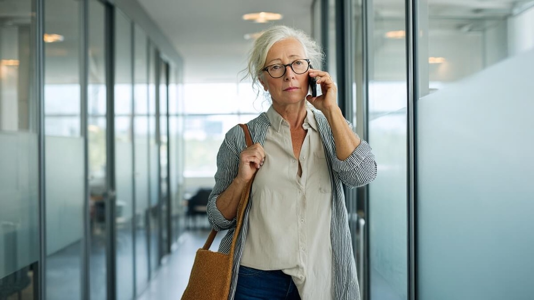 Une femme de 55 ans au téléphone dans un couloir de bureau, entre deux obligations © SeniorActu Une femme de 55 ans au téléphone dans un couloir de bureau, entre deux obligations © SeniorActu