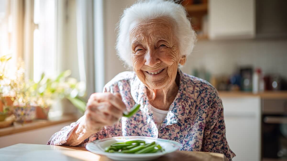 Une centenaire souriante mange des haricots verts dans sa cuisine © SeniorActu